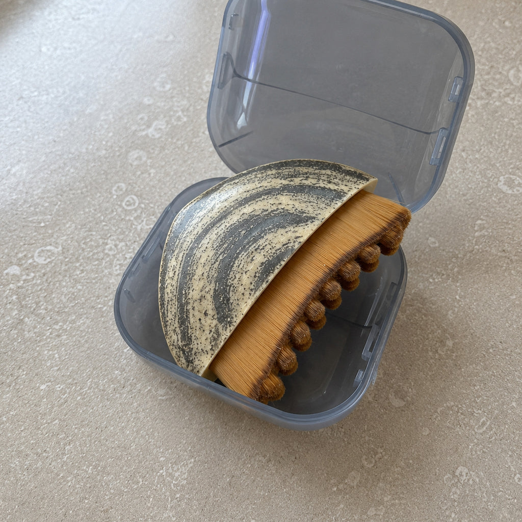 The Cueva Contour Brush sitting in its hinged grey storage case on a bathroom counter