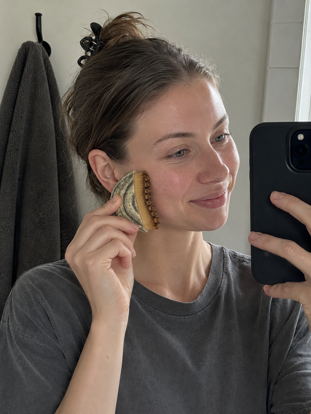 Woman smiling at her reflection in a bathroom mirror while holding the Cueva Contour Brush against her cheekbone